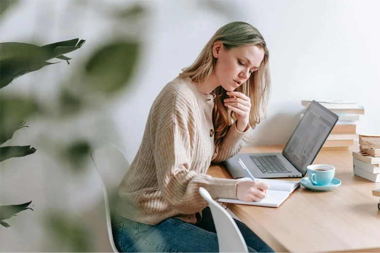 Mujer en un entorno de teletrabajo, sentada en un escritorio con un ordenador portátil y un cuaderno, realizando sus actividades laborales