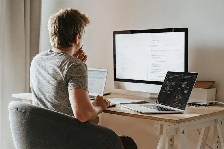 Hombre trabajando en teletrabajo, sentado en un escritorio con dos laptops y una pantalla de computadora