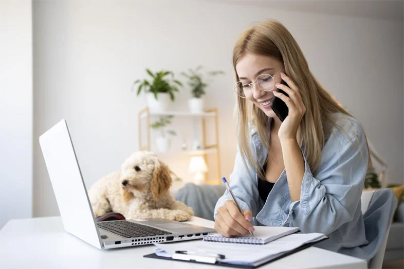 Mujer hablando por teléfono con un perro sentado frente a su laptop, ilustrando las ventajas del teletrabajo